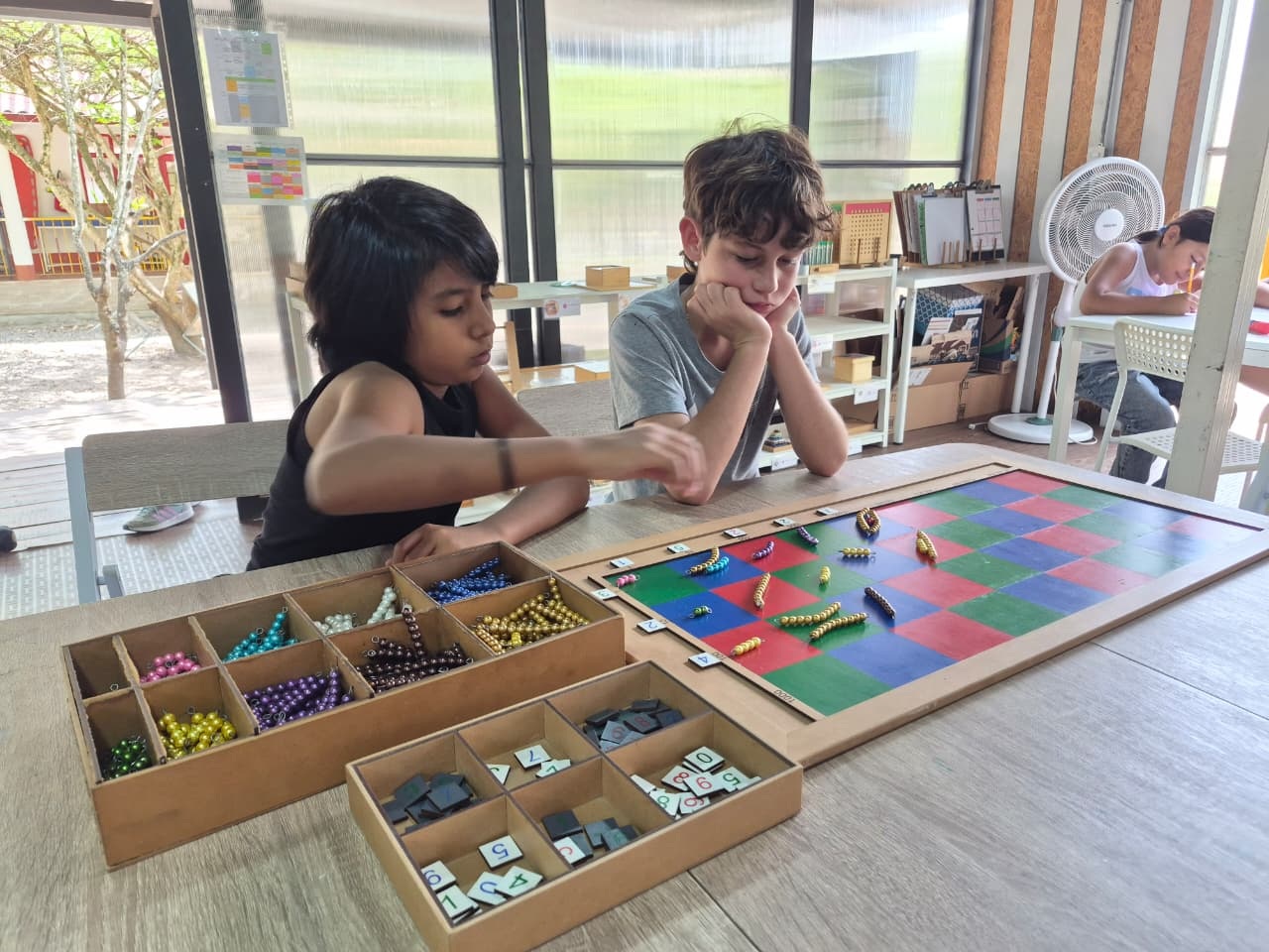 Dos estudiantes trabajan juntos con material Montessori de matemáticas en un ambiente preparado.