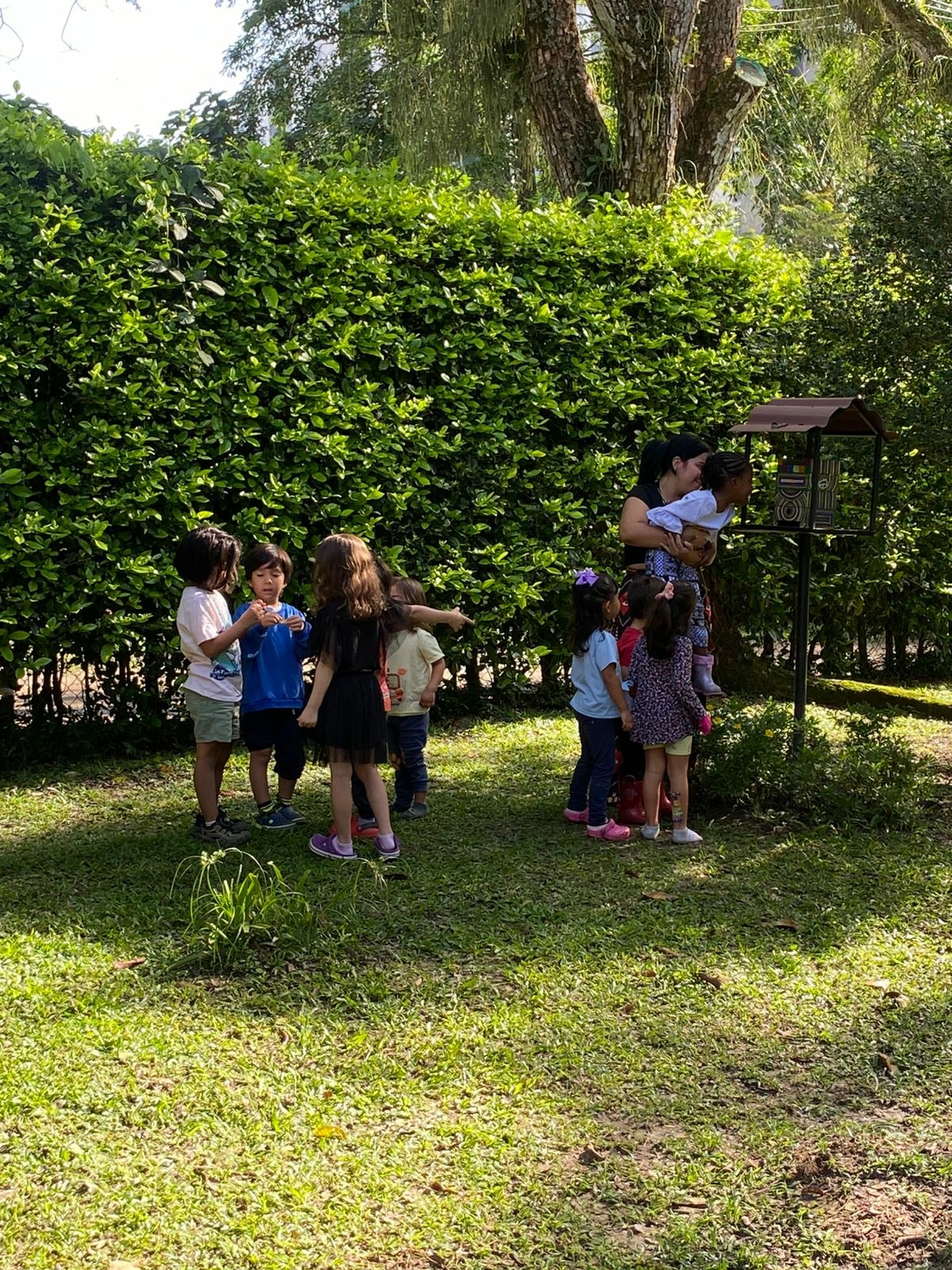 Una persona adulta sostiene a una niña pequeña mientras otras niñas y niños se encuentran en el jardín.